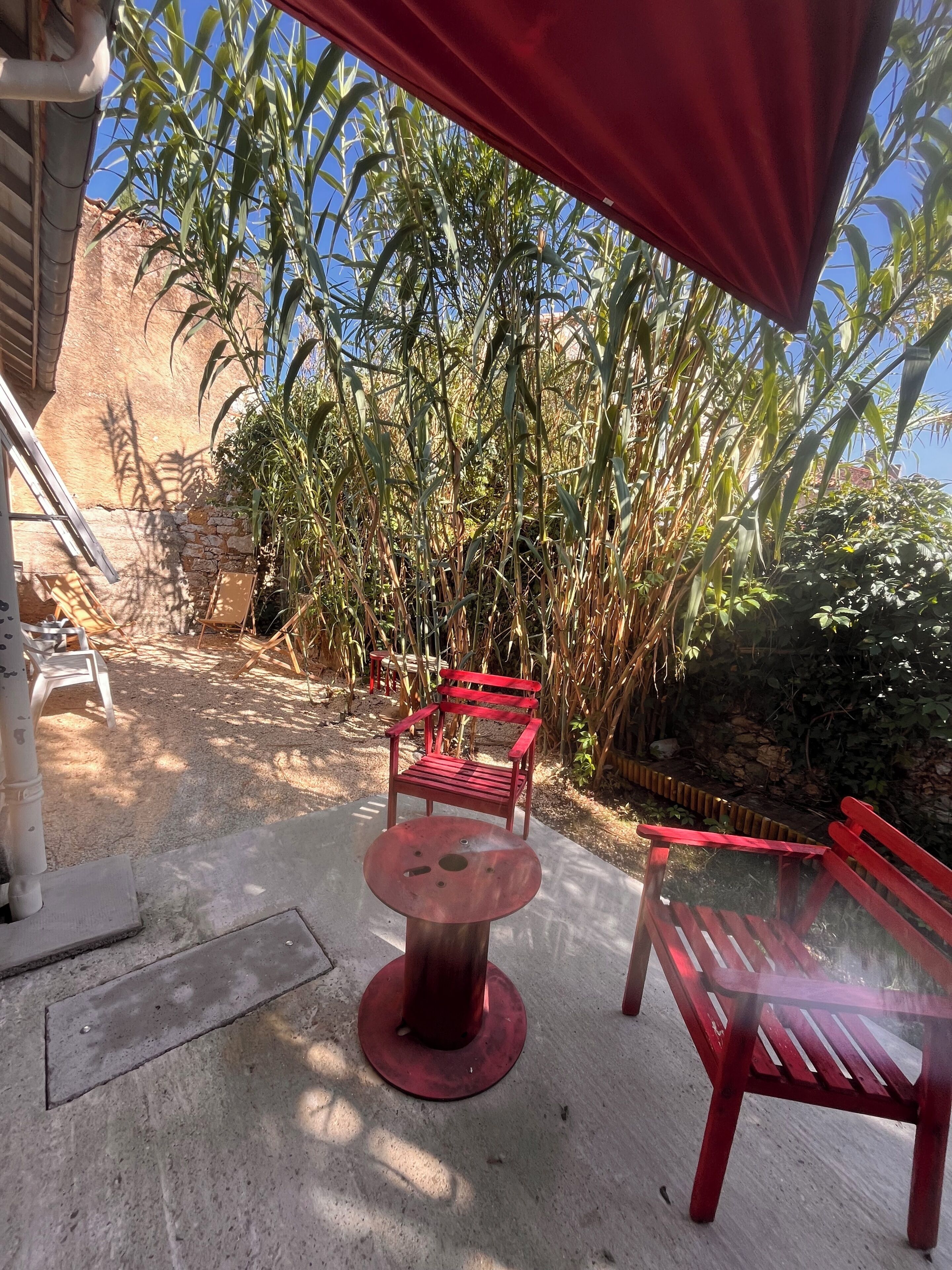 An outdoor seating area with a round table and two red chairs, surrounded by tall plants and a stone wall.