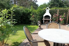 An outdoor patio with a round table and two wicker chairs, a white barbecue grill, and a lush garden with various plants and flowers. - Holiday home in Prerow with swimming pool (Prerow)