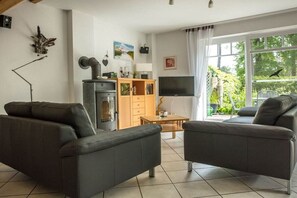 A modern living room with two sofas, a television, a fireplace, and a large window with a view of greenery. - Holiday home in Prerow with swimming pool (Prerow)