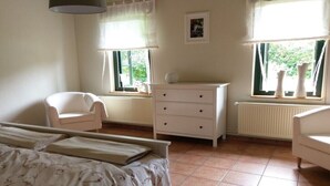 Ein Schlafzimmer mit einem Bett, einem Sessel, einer Kommode und zwei Fenstern mit Vorhängen.