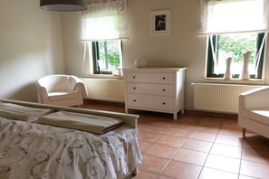 Un dormitorio con cama, una silla, cómoda y dos ventanas con cortinas.