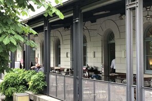Ein Straßencafé mit Tischen und Stühlen, umgeben von einem Metallzaun und Pflanzen.