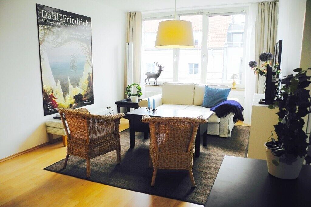 A living room with a sofa, two wicker chairs, a coffee table, a poster on the wall, and a window with a view of a building and a statue.