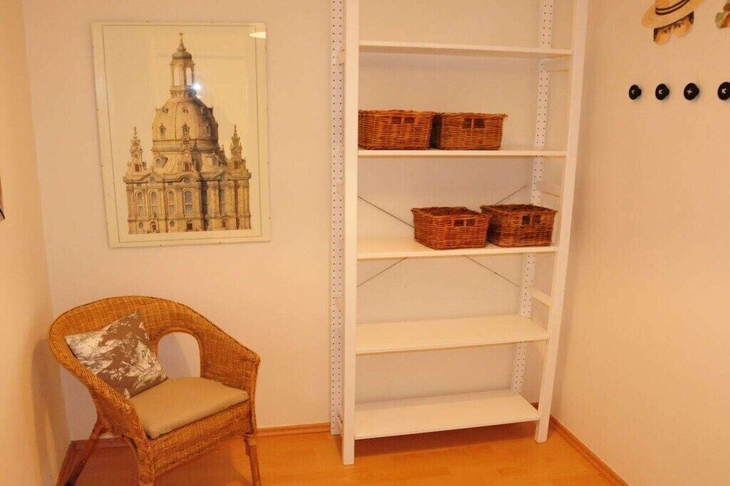 Ein Zimmer mit einem Korbstuhl, einem weißen Regal mit Körben und einem gerahmten Bild eines Gebäudes.