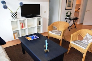 Ein Wohnzimmer mit einem Flachbildfernseher auf einem weißen Regal, zwei Korbstühlen, einem dunklen Couchtisch mit einer Kerze und einem Bücherregal.