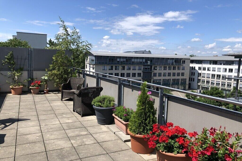 A rooftop terrace with potted plants, outdoor furniture, and a view of surrounding buildings.
