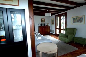 Ein Wohnzimmer mit einer Couch, einem grünen Sessel, einem runden Tisch und einer Kommode mit gerahmten Bildern.