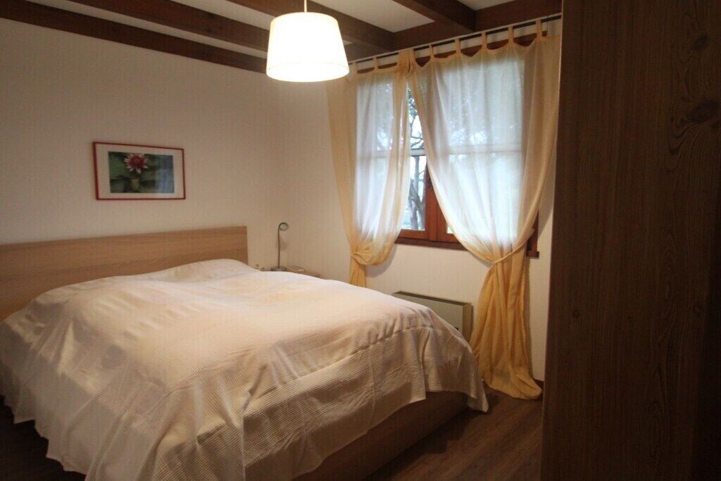 Un dormitorio con cama, mesita de noche, lámpara, ventana con cortinas e imagen enmarcada en la pared.