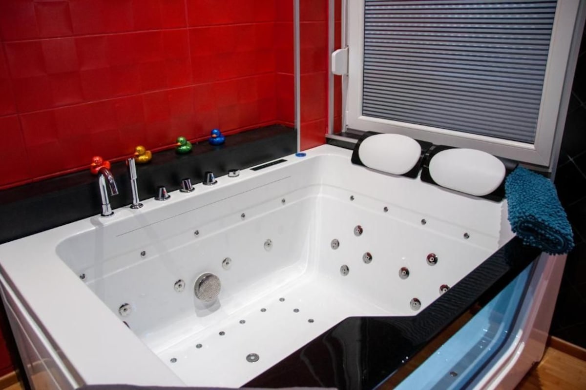 A modern bathroom with a white bathtub, a red tiled wall, and a window with blinds.