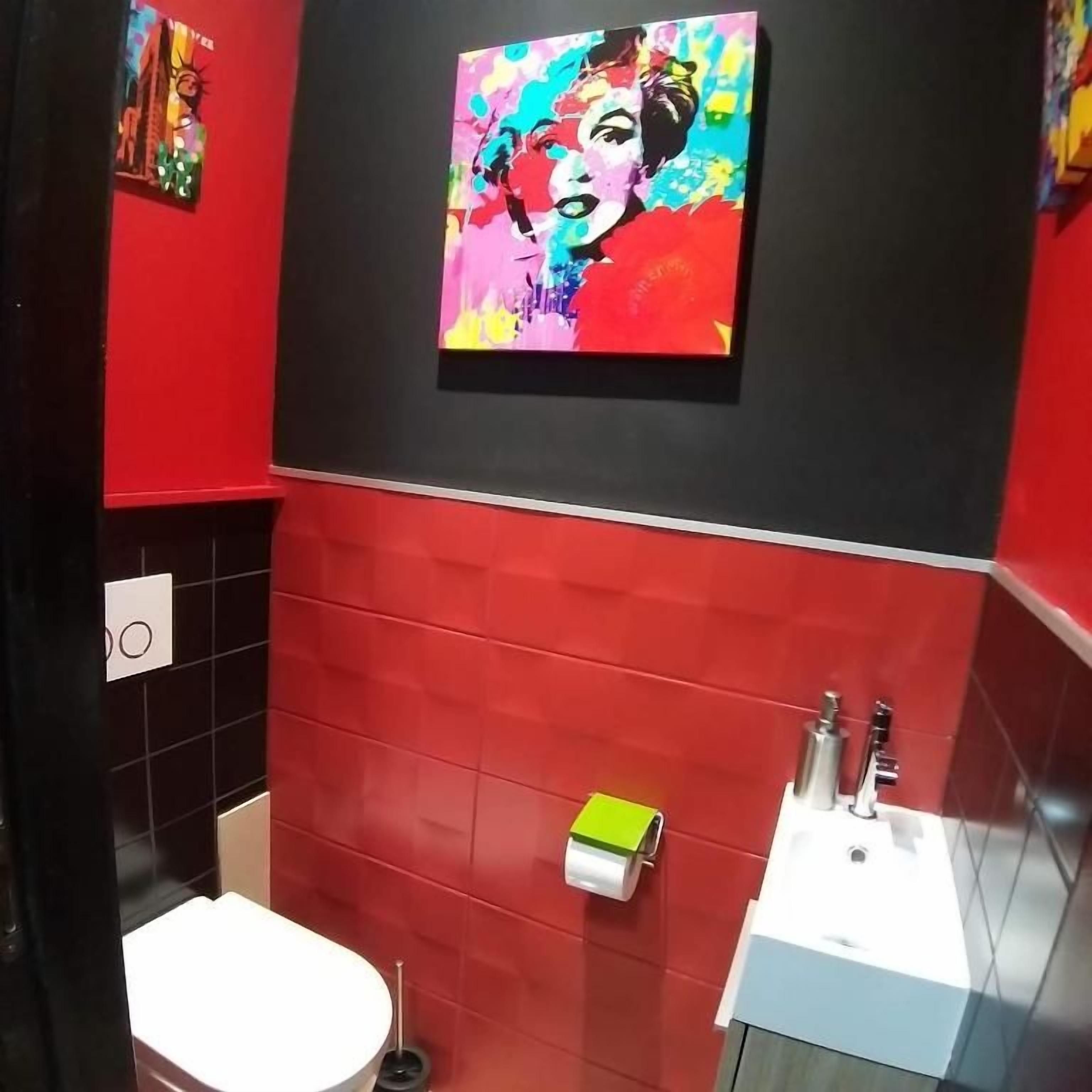 Een badkamer met rode betegelde wanden, een wit toilet, een wastafel met spiegel en een kleurrijk schilderij boven de wastafel.