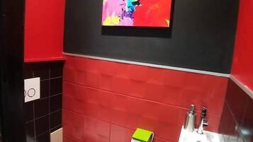 Een badkamer met rode betegelde wanden, een wit toilet, een wastafel met spiegel en een kleurrijk schilderij boven de wastafel.