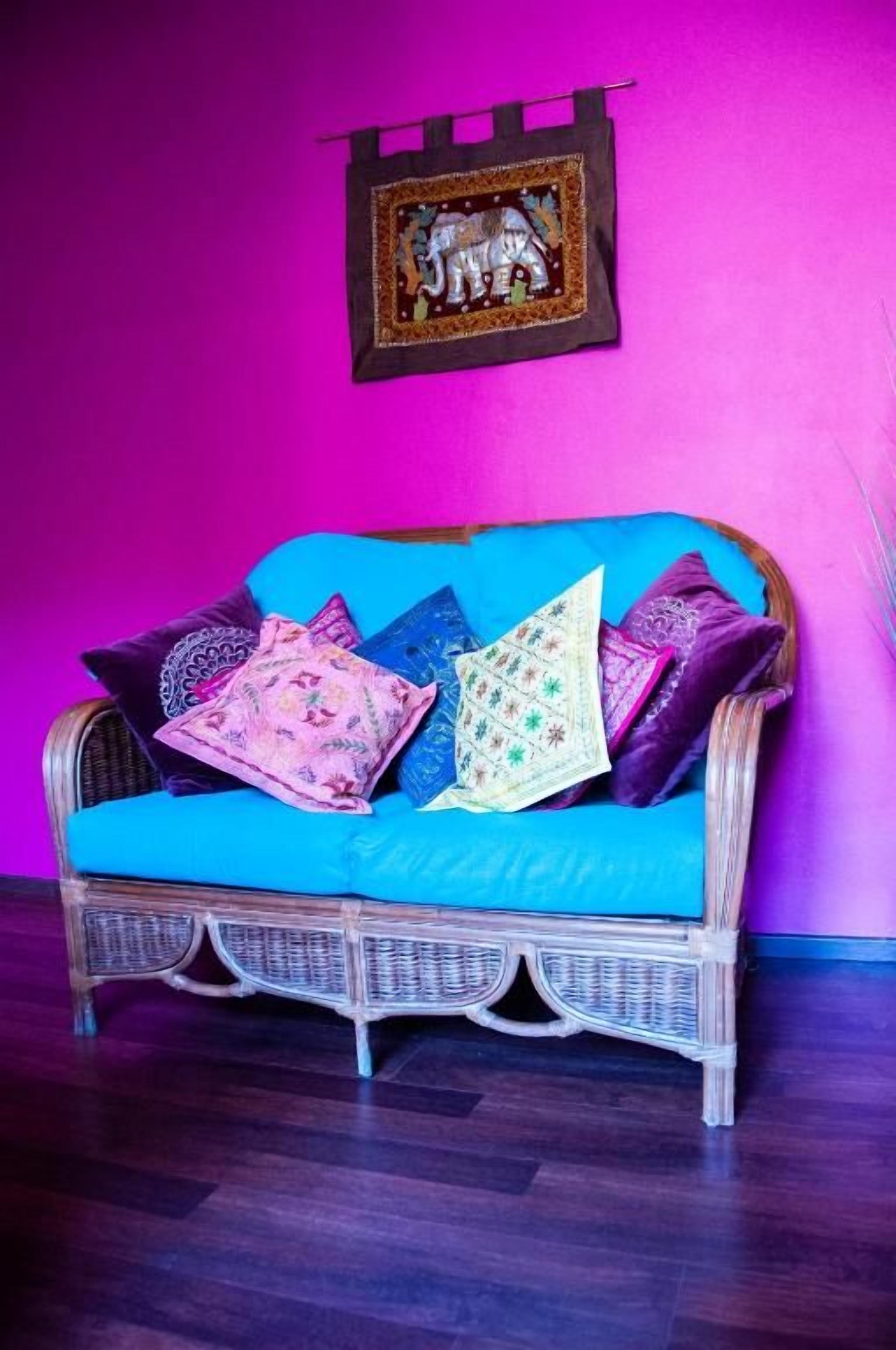 Un sofá de mimbre con un cojín azul frente a una pared rosa, junto con un cuadro con la imagen de un elefante.