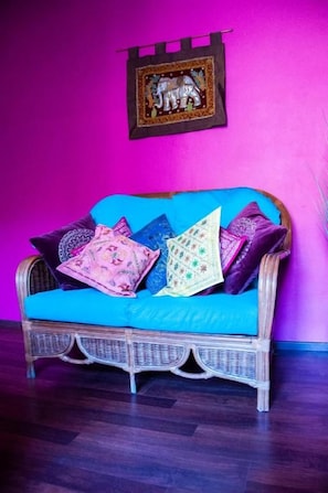 Un sofá de mimbre con un cojín azul frente a una pared rosa, junto con un cuadro con la imagen de un elefante.
