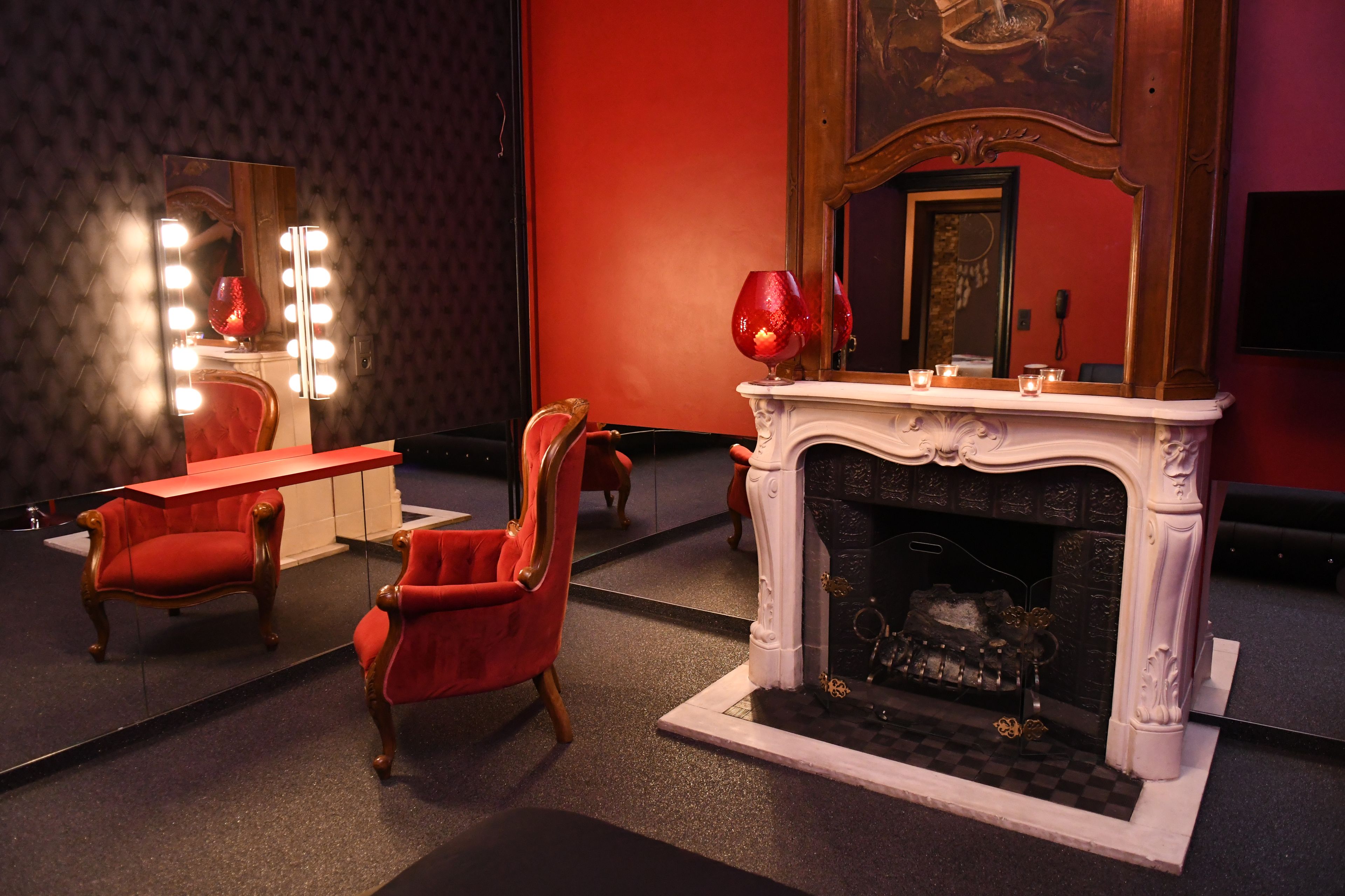 A room with a mirror, vanity, red chairs, a fireplace, and a red wall.