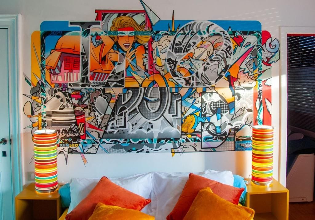 A room with a colorful graffiti wall, a bed with orange pillows, and two striped lamps on side tables.