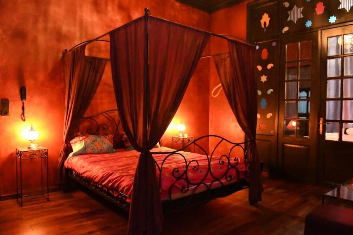 A four-poster canopy bed with a red bedspread, two bedside lamps, and a wooden floor.