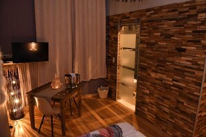 A room with a wooden floor, a dining table with chairs, a TV mounted on the wall, and a sliding glass door leading to a bathroom. - Vip room 209 (Trooz)