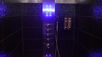 Ducha moderna con panel vertical y múltiples rociadores, frente a una pared de azulejos oscuros con textura.
