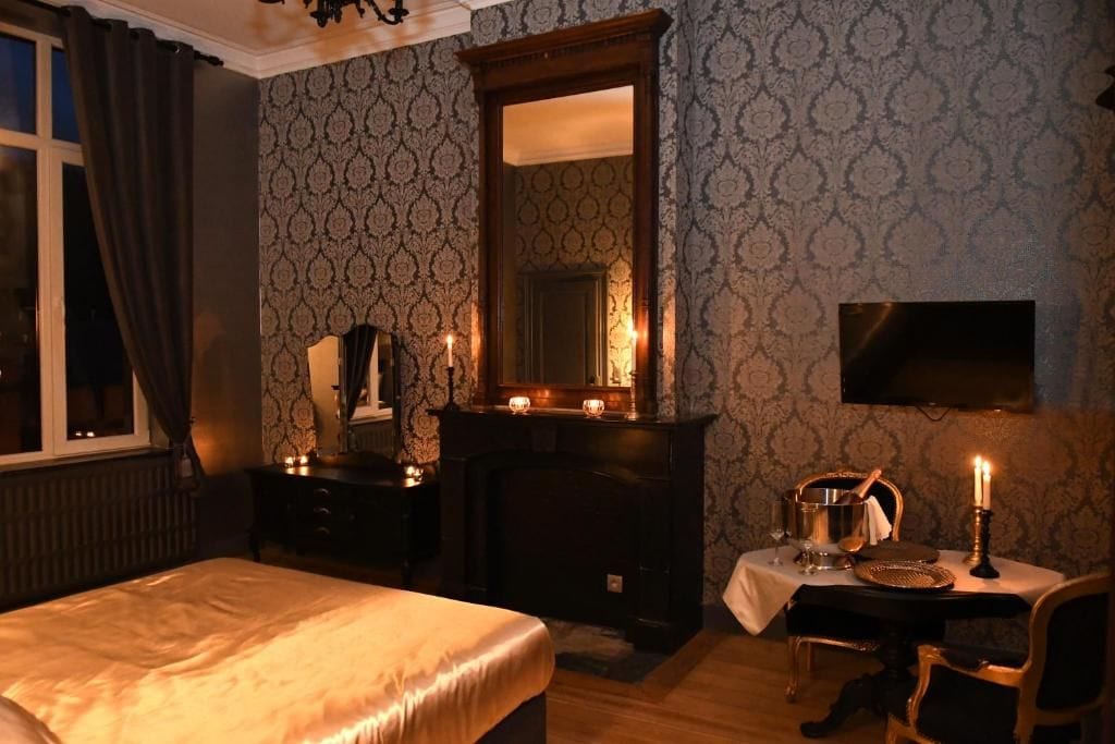 A hotel room with a bed, a desk with a chair, a fireplace, a mirror, and a television mounted on the wall.