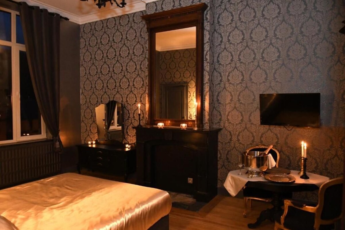 Una habitación de hotel con una cama, un escritorio con silla, chimenea, espejo y televisión montada en la pared.