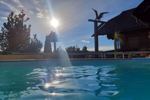 A swimming pool with clear water, surrounded by trees and a building with a thatched roof. - Kraxlhütte Carinthia (Dröschitz)