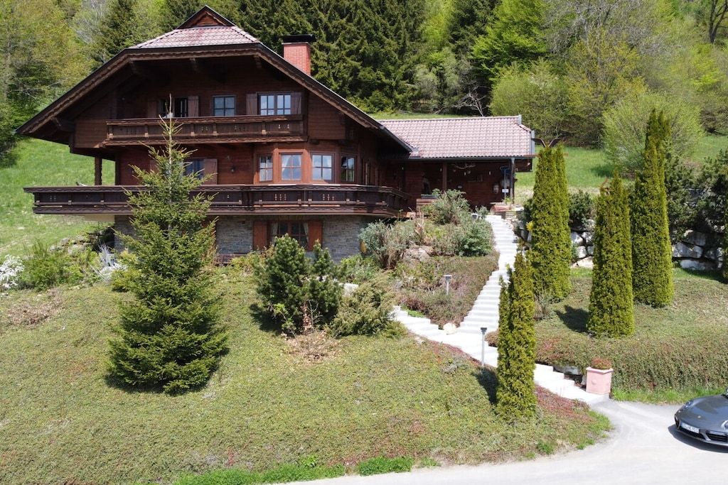 Un chalet en bois doté d’un balcon, entouré d’un jardin bien entretenu et accessible par un escalier en pierre menant à l’entrée.