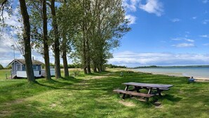Un espace verdoyant avec une table de pique-nique, des arbres, une petite cabane et une étendue d’eau en arrière-plan.