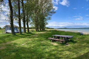 Een grasveld met een picknicktafel, bomen, een klein huisje en een stuk water op de achtergrond.