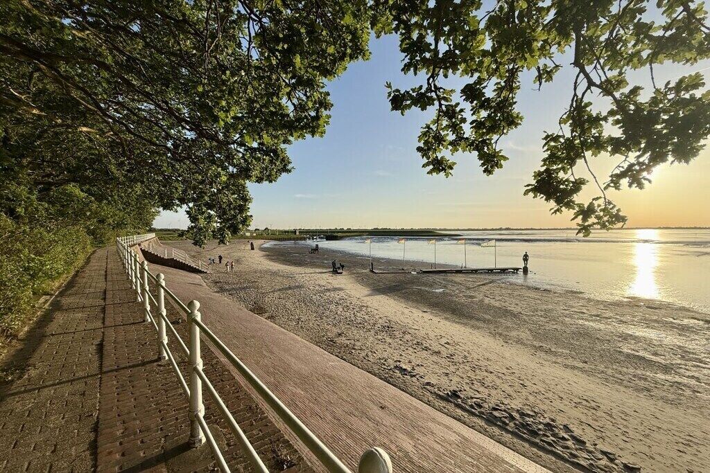 En asfalterad gångväg leder till en strand med ett räcke, en klar himmel och solen som går ner över vattnet.