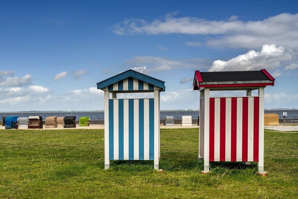 Deux cabanes de plage aux motifs rayés, l’une bleue et l’autre rouge, se dressent sur un champ verdoyant, offrant une vue sur la mer.