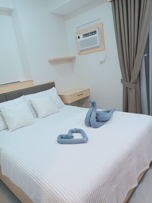Ett hotellrum med en säng, kuddar, ett nattduksbord, ett litet bord, en väggmonterad luftkonditioneringsanläggning och ett fönster med gardiner.