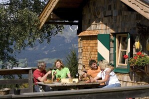Eine Holzhütte mit einem für vier Personen gedeckten Esstisch, umgeben von viel Grün und Bergen im Hintergrund.