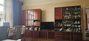 A room with a wooden cabinet containing various items, a television, and a window with curtains. - Enjoyable 3-bedroom house with AC in charming Gyumri (Gyumri)