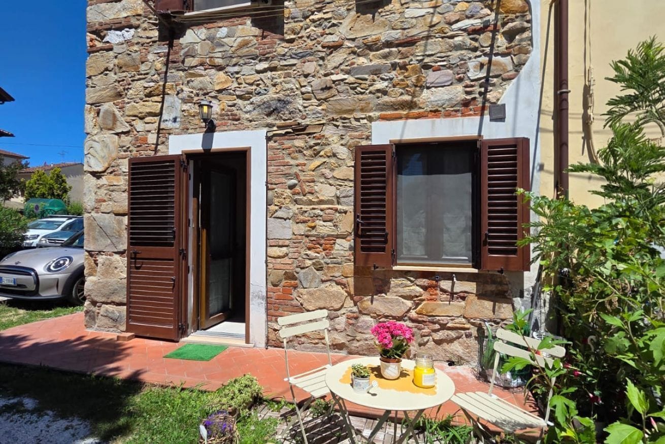 A stone building with two open doors and windows with shutters, a small table with a vase of flowers, and a bench.