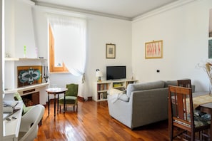 Ein Wohnzimmer mit einem grauen Sofa, einem Fernseher, einem Bücherregal und einem Fenster mit Vorhängen.