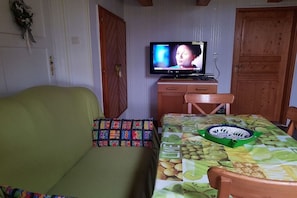 A room with a television on a wooden cabinet, a green sofa with colorful pillows, and a table with a fruit-patterned tablecloth. - Holiday apartment in Dunum with swimming pool (Dunum)
