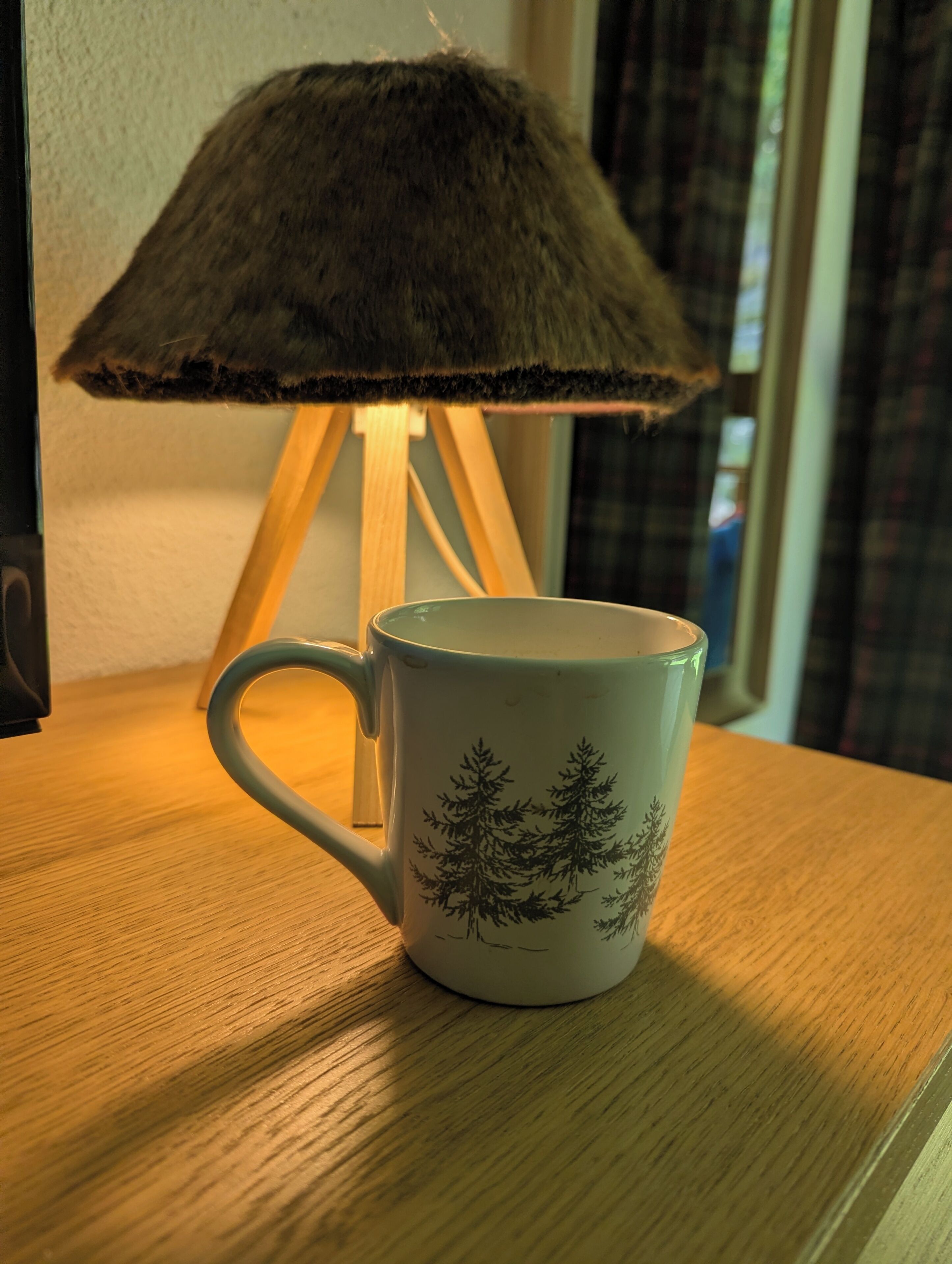 Una lámpara con pantalla de textura suave y una taza con diseño de árbol sobre una superficie de madera.