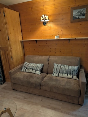Een kamer met houten lambrisering, een bruine bank, twee kussens met dessin, een wandlamp en een lijst met een afbeelding.
