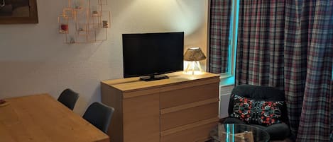 A living room with a wooden TV stand, a glass table, a sofa with a patterned cushion, and a plaid curtain.