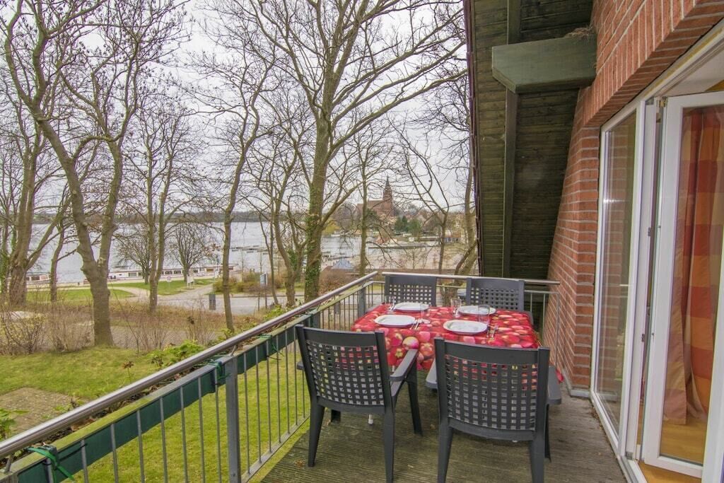 Un balcon extérieur avec une table dressée pour le dîner, offrant une vue sur une étendue d’eau et des arbres.