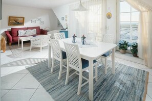 Ein modernes Wohnzimmer mit einem weißen Sofa, einem Esstisch mit Stühlen und einem großen Fenster mit Vorhängen.
