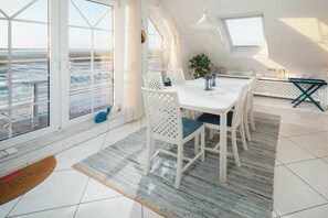 Ein Essbereich mit einem weißen Tisch und Stühlen, Blick durch große Fenster und eine kleine Pflanze auf dem Tisch.