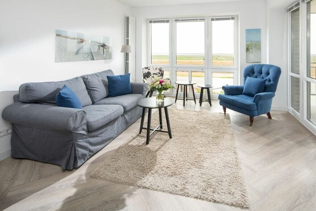 Una sala de estar con un sofá gris, un sillón azul, una mesita redonda con un jarrón y un ventanal con vistas.