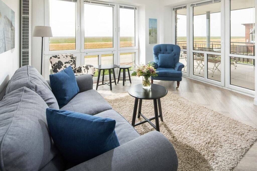 A modern living room with a sofa, a blue armchair, and a small table with a vase. Large windows provide a view of the outdoors.