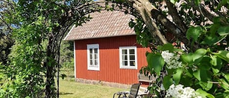 A red wooden house with white windows, surrounded by greenery and a bench.