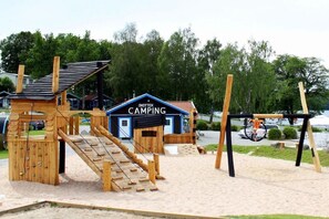 A wooden play structure with a climbing wall and a slide, a small blue building labeled 'SKOTTEK CAMPING', and a swing set. - directly on the lake in Sweden (Ulricehamn)