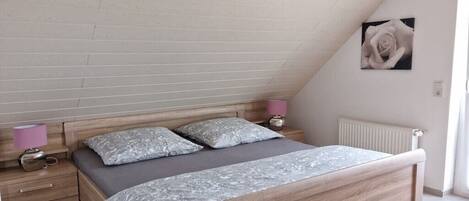 Ein Schlafzimmer mit schrägverdeckter Decke, einem Holzbett, zwei Nachttischen mit Lampen und einem an der Wand hängenden Bild.