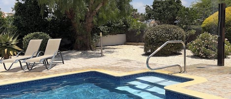 A swimming pool with a tiled edge, surrounded by lounge chairs and a tree.