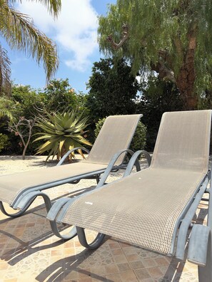Two lounge chairs with metal frames and woven seats, positioned on a tiled patio with a palm plant and a large tree in the background. - Family Villa With Large Pool, Home Away From Home (Pegeia)