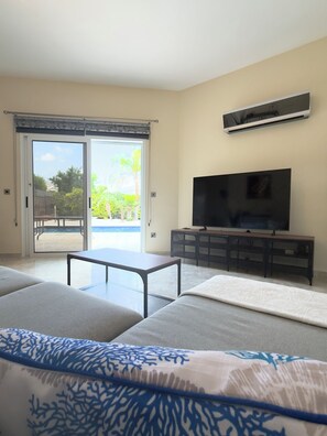 A living room with a sofa, a coffee table, a TV on a stand, and a sliding glass door leading to an outdoor area. - Family Villa With Large Pool, Home Away From Home (Pegeia)